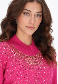 Felroze trui met een ronde halslijn, versierd met verspreide gouden pailletten op de borst, met ribgebreide manchetten en onderrand.