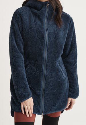 Frau trägt eine marineblaue Fleecejacke mit durchgehendem Reißverschluss, hohem Kragen, Fronttaschen und langen Ärmeln.