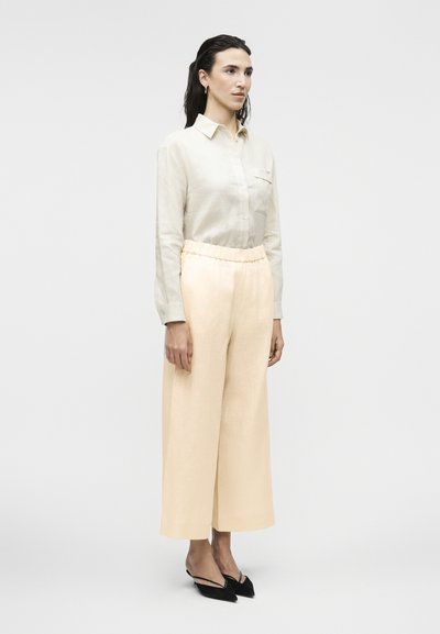 Femeie în picioare, purtând o cămașă bej deschis cu nasturi, pantaloni crem cu picioare largi, cercei aurii rotunzi și pantofi negri cu toc ascuțit și baretă slingback.