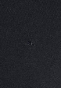 Czarna bawełniana tkanina o prążkowanej fakturze. Blisko dolnego środka wyhaftowano niewielkie, ton w ton logo, zachowując minimalistyczny design.