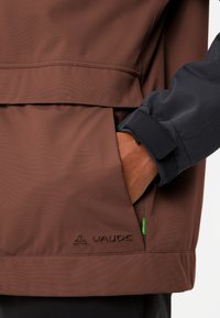 Giacca impermeabile marrone con manica nera, dotata di tasca con zip, polsini regolabili e logo Vaude ricamato all'orlo.