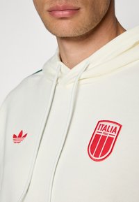 Cream hoodie with white drawstrings, featuring red embroidered Adidas logo and Italy national team crest on the chest. Soft texture.