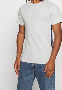 Muž v světle šedém tričku s krátkým rukávem a malým logem na hrudi, doplněný modrými denimovými džíny s námořnicko-modrými a bílými postranními pruhy.