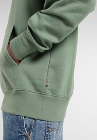 Närbild av en persons arm klädd i en grön hoodie med en framficka, tillsammans med blå jeans. En logotyp är synlig på hoodien.