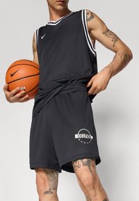 Maillot de basket noir et shorts en maille, avec des accents blancs et un logo "CELTICS" sur les shorts. Tenant un ballon de basket.