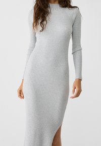 Abito in maglia grigia a costine con maniche lunghe e colletto alto. Presenta una fessura laterale e una silhouette aderente. Tessuto morbido e materiale elasticizzato.