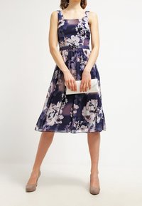 Žena ve bezramenných námořnických květinových šatech s páskem, držící bílou kabelku, obutá v béžových vysokých podpatcích, stojící proti jednobarevnému světlému pozadí.