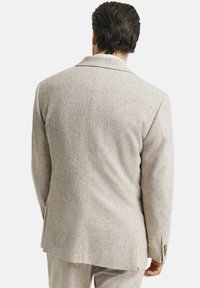 Mann mit dunklem Haar, von hinten gesehen, trägt einen hellbeigen, gesprenkelten Sakkos und passende Hosen vor einem einfarbigen Hintergrund.