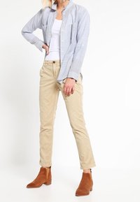 Chemise rayée bleu clair et blanche, débardeur blanc, pantalon beige et bottines brunes à la cheville. Présente des poignets à boutons et un design à jambe droite.