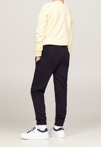 Person, der schwarze Jogginghosen, weiße Sneaker mit navy-farbenen Details und einen cremefarbenen Hoodie trägt, steht mit den Händen in den Taschen und blickt weg.