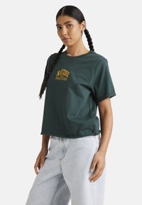 Green cotton t-shirt with short sleeves, round neck, and yellow embroidered logo. Relaxed fit and stitched hem. Worn with light denim jeans.
