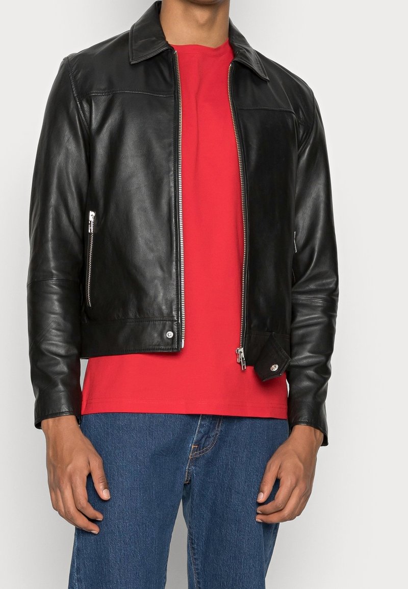 Uomo che indossa una giacca di pelle nera sopra una camicia rossa e jeans blu, in piedi con le mani rilassate lungo i fianchi.