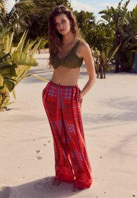 Grüner gestrickter Bralette mit Drehdetail, kombiniert mit lockeren roten Palazzo-Hosen mit Mustern aus Wirbeln, Zickzacken und Punkten. Sandiger Strand als Hintergrund.