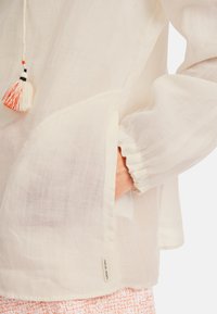 Leichte weiße Bluse mit gerafften Ärmeln und einer Seitentasche. Verfügt über eine dekorative Quaste an einem Kordelzug am Ausschnitt. Strukturierter Stoff.