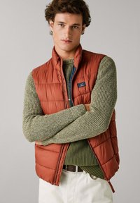Gilet imbottito arancione sopra un maglione verde testurizzato. Il gilet presenta una cerniera e dettagli con logo, indossato con pantaloni chiari. Sfondo semplice.