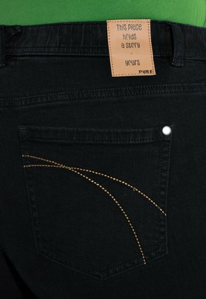 Nærbillede af baglomme på sorte jeans med buet beige syning og tan-mærke med teksten "Dette stykke rummer en historie - din" mod en grøn skjorte.