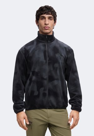 Hombre de pie mirando hacia adelante con una chaqueta de forro polar negra con cremallera y pantalones verde oliva sobre un fondo liso.