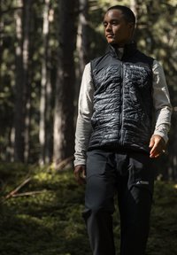 Schwarze isolierte Weste über einem hellen Langarmshirt, kombiniert mit dunklen Hosen mit dem 'Terrex'-Logo. Waldhintergrund, natürliches Licht.