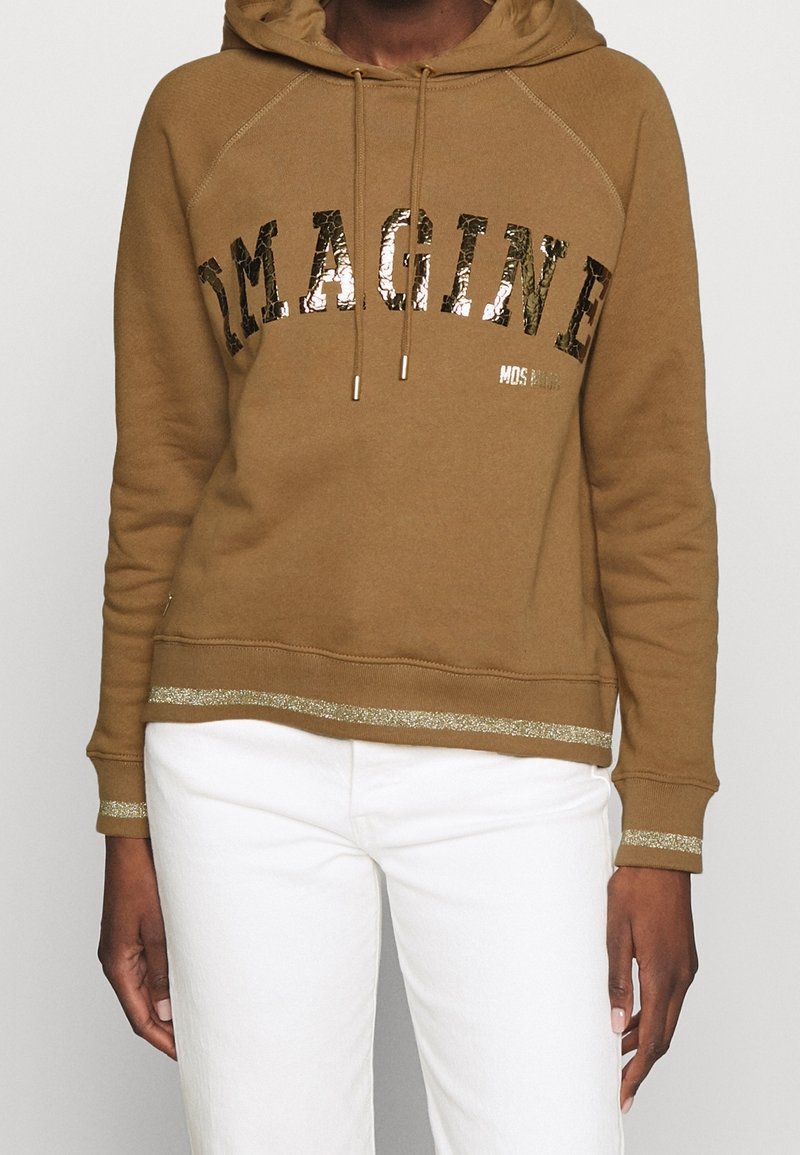 Personne portant un sweat à capuche marron avec "IMAGINE" en lettres brillantes et un pantalon blanc, avec un bord pailleté doré aux poignets et à l'ourlet du sweat.