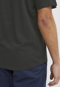 Melna kokvilnas T-kreklu ar noapaļotu apakšmali, īsām piedurknēm un gludu tekstūru, kombinē ar tumši ziliem biksēm, redzama daļa no rokas.