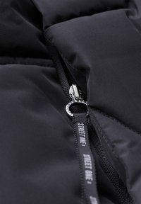 Svart puffjacka sektion med en framträdande silverdragkedja. Har stickade sömmar och en ögla för dragkedjan med texten "STREET ONE".