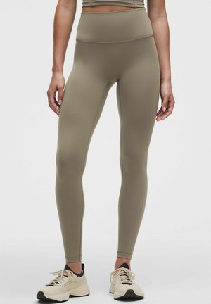 Donna che indossa leggings taupe a vita alta e sneakers crema in piedi davanti a uno sfondo chiaro e uniforme.