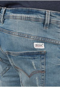 Acercamiento del bolsillo trasero de unos pantalones de mezclilla azul claro con etiqueta de marca visible y sutiles detalles desgastados.