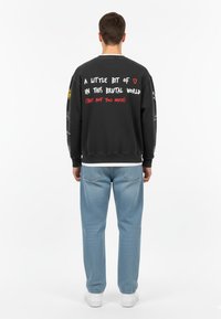 Felpa nera con testo colorato sul retro; presenta un colletto girocollo, vestibilità comoda e polsini a costine. Indossata con jeans azzurri e sneaker bianche.