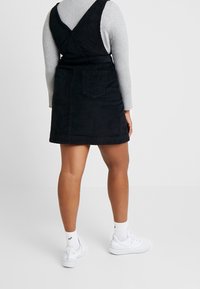 Schwarzes Kord-Mischkleid mit tailliertem Design, featuring einem V-Ausschnitt hinten und einer Rückentasche. Getragen über einem grauen, gerippten Langarmoberteil und weißen Sneakers.