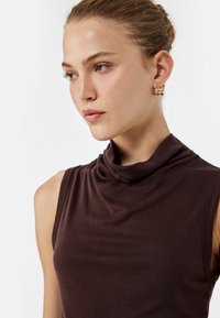 Brązowy bezrękawnik z dekoltem typu cowl. Wykonany z prążkowanej tkaniny, o gładkiej fakturze i subtelnym połysku. Widoczne złote kolczyki.