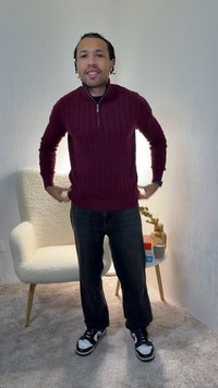 Μπορντό πουλόβερ με πλέξη καλώδιο και φερμουάρ στο γιακά, συνδυασμένο με σκούρο τζιν παντελόνι και ασπρόμαυρα αθλητικά παπούτσια, στέκεται σε ένα χώρο με απαλή φωτισμό.