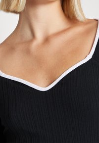 Zwarte geribbelde top met een brede V-halslijn met witte rand, die de gestructureerde stof en schone lijnen benadrukt zonder patronen of versieringen.