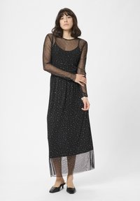 Rochie maxi neagră cu un model de puncte argintii, având un overlay transparent cu mâneci lungi, corp ajustat și o siluetă evazată.