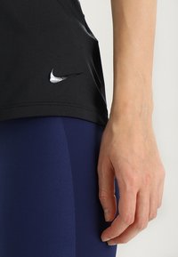 Sort træningstop med et Nike-logo på nederste venstre side, parret med blå leggings. Stoffet ser glat og tætsiddende ud.