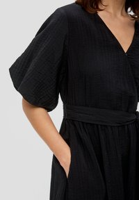 Abito nero con texture, maniche corte e ampie, scollatura a V e vita con cintura. Presenta tasche laterali e tessuti sovrapposti per una maggiore dimensione.