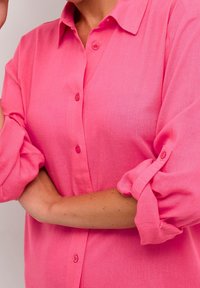Różowa koszula z długim rękawem w lekkim materiale, z klasycznym kołnierzykiem, podwiniętymi rękawami oraz gładką fakturą i minimalną ilością detali.