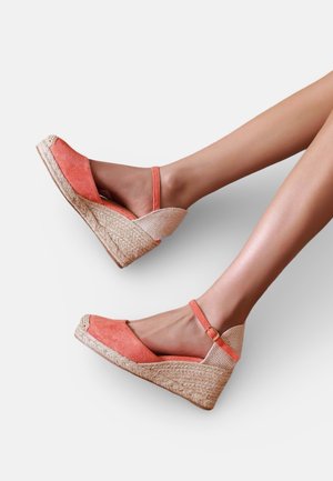 Jambes portant des sandales compensées espadrilles corail avec brides à la cheville, avec des semelles plateformes en jute tressé sur fond blanc.