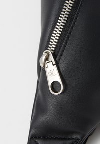 Zwarte leren tas sectie met een zilveren rits en een ronde trekker met gegraveerd 'CK' logo. Gladde textuur met zichtbare stiksel details.