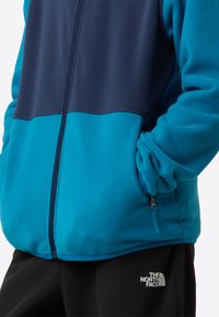 Fleecejacka med färgblock i blått och marinblått, hel dragkedja och ficka med blixtlås, kombinerad med svarta byxor med en logotyp.