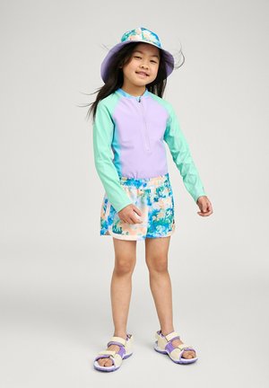 Junges Mädchen lächelt, trägt ein langärmeliges Schwimmshirt in Pastell-Lila und Minzgrün, geblümte Shorts, einen Bucket-Hut und cremefarbene Sandalen mit lila Sohlen.