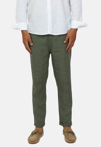 Pantaloni in lino khaki con una vestibilità slim, orli risvoltati e una texture liscia; abbinati a una camicia bianca a maniche lunghe con bottoni e scarpe chiare.