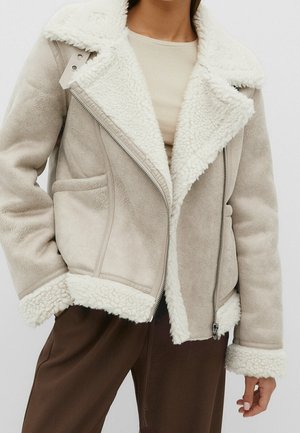 Gros plan d’une personne portant une veste beige en shearling avec une doublure en polaire blanche, une chemise beige et un pantalon marron, debout, une main dans la poche.