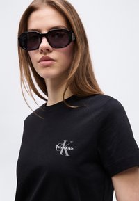 Νεαρή γυναίκα που φοράει μαύρο t-shirt Calvin Klein και μεγάλα μαύρα γυαλιά ηλίου, κοιτάζοντας μπροστά με ουδέτερη έκφραση σε απλό φόντο.