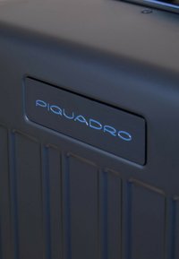 Schwarzer Hartschalenkoffer mit vertikal geriffelter Oberfläche. Verfügt über ein silbernes Logo mit dem Schriftzug "PIQUADRO" in moderner Schriftart. Glatte Oberfläche.