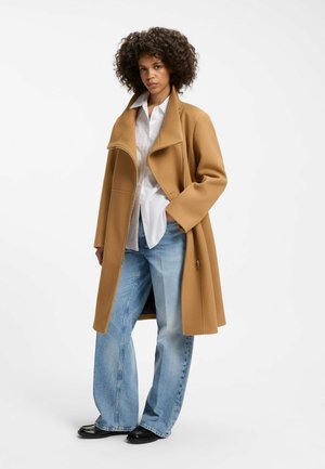 Donna con capelli ricci che indossa un cappotto color cammello, una camicia bianca, jeans larghi di colore azzurro chiaro e scarpe nere, in piedi con una mano nella tasca.