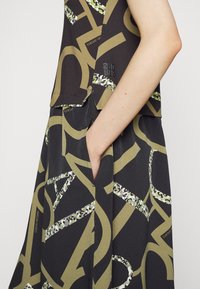 Ärmelloses Oberteil aus dunklem Stoff, mit großen, fetten Buchstaben in Olivgrün und grünen Musterakzenten. Verfügt über eine Seitentasche und ein Logodetail.