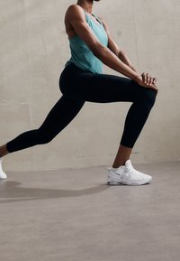 Persoon in zwarte leggings, teal tanktop en witte sneakers voert een voorwaartse lungestretch uit tegen een beige muur.