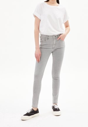 Kvinde iført hvid kortærmet t-shirt, lysegrå stramme jeans og sort-hvide sneakers, stående med den ene hånd i lommen.