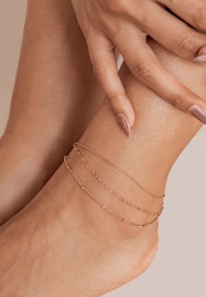 Gold ankle chains in various designs, featuring delicate links and a layered appearance, worn on a tanned ankle with smooth skin.