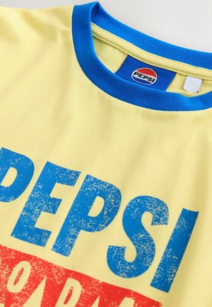 Żółta koszulka z niebieskim okrągłym dekoltem, z dużym, niebiesko-czerwonym postarzałym napisem "PEPSI". Wewnątrz kołnierza widoczna metka.